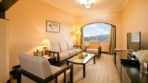 A hotel room with a sofa, armchair, and a dining table. There is a city view through a large window, a chandelier, and a framed picture on the wall. - Yindo Jasper Hotel Zhuhai (Zhuhai)