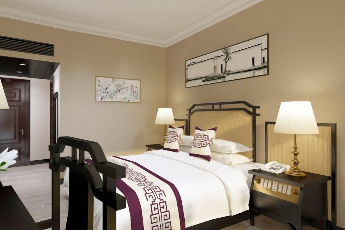 Habitación de hotel con cama, dos mesitas de noche con lámparas, un reloj de pared y un cuadro.
