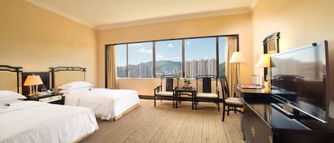 A hotel room with two beds, a desk, a TV, and a large window offering a city view.