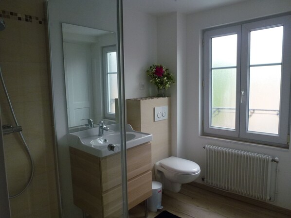 Une salle de bain moderne dotée d’une douche à parois de verre, d’un lavabo, de toilettes et d’une fenêtre laissant entrer la lumière naturelle.