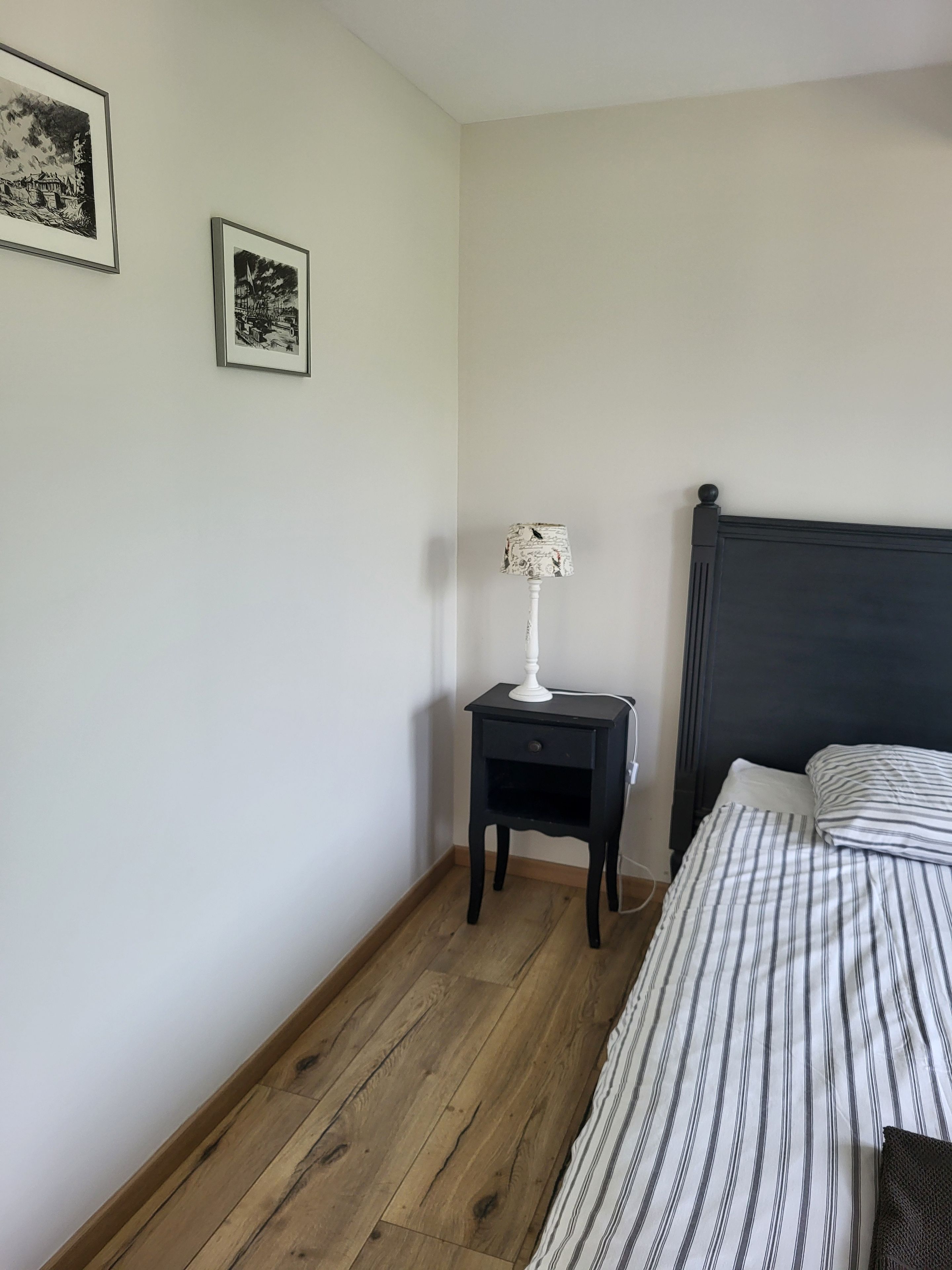 Quarto com cama, mesa de cabeceira com abajur e duas fotografias emolduradas na parede.