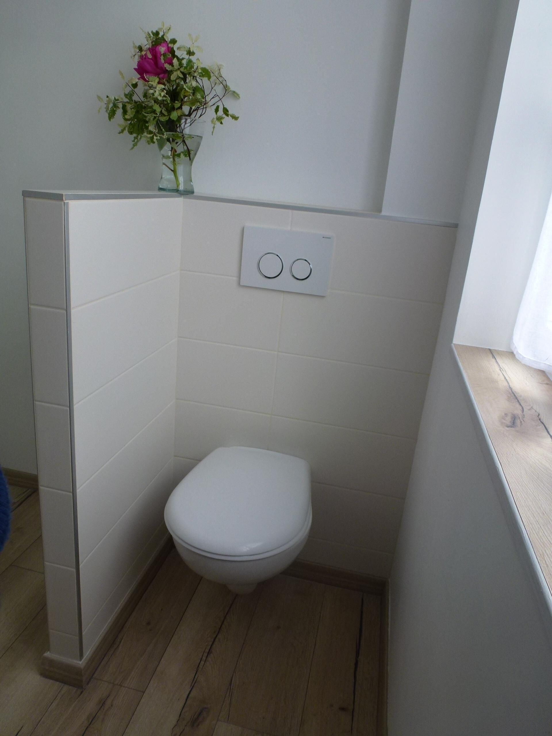 Een kleine badkamer met een wit toilet, een in de muur gemonteerde lichtschakelaar en een bloempot met roze bloemen.