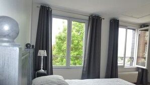 Une chambre à coucher avec un lit, deux fenêtres avec des rideaux, un radiateur et une table de chevet avec une lampe.