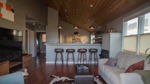 A modern living room with a large flat-screen TV, a sofa, a coffee table, and a cowhide rug. There are wooden floors, a wooden ceiling, and a kitchen area with a bar stool and a refrigerator.