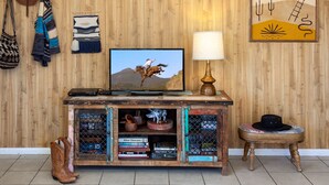 Una sala rústica con pared de madera, cráneo de vaca montado, televisor, lámpara, estantería y botas de vaquero.