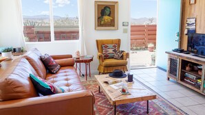 Sala de estar con un sofá de cuero marrón, una mesa de centro de madera y un sillón con un cojín colorido. Hay un cuadro en la pared y una puerta que da a un espacio exterior.