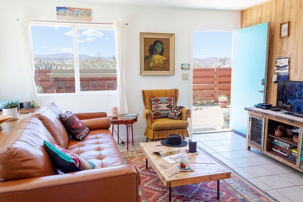 Ein Wohnzimmer mit braunem Ledersofa, einem Holztisch und einem Sessel mit farbenfroher Kissen. Ein Gemälde hängt an der Wand und eine Tür führt in einen Außenbereich.