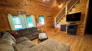 A cozy log cabin interior with a sofa, a coffee table, a TV, and a staircase leading to a loft area. - Creekside Log Cabin, Ideal Family Vacation! Hot tub, Fire Pit, Tubing, Fishing (Robbinsville)