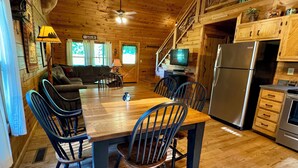 A wooden cabin interior with a dining table, chairs, a sofa, a television, and a refrigerator. - Creekside Log Cabin, Ideal Family Vacation! Hot tub, Fire Pit, Tubing, Fishing (Robbinsville)