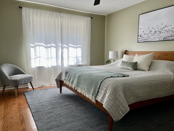 Un dormitorio con cama, una silla, una alfombra, una ventana con cortinas y una lámpara fijada a la pared.