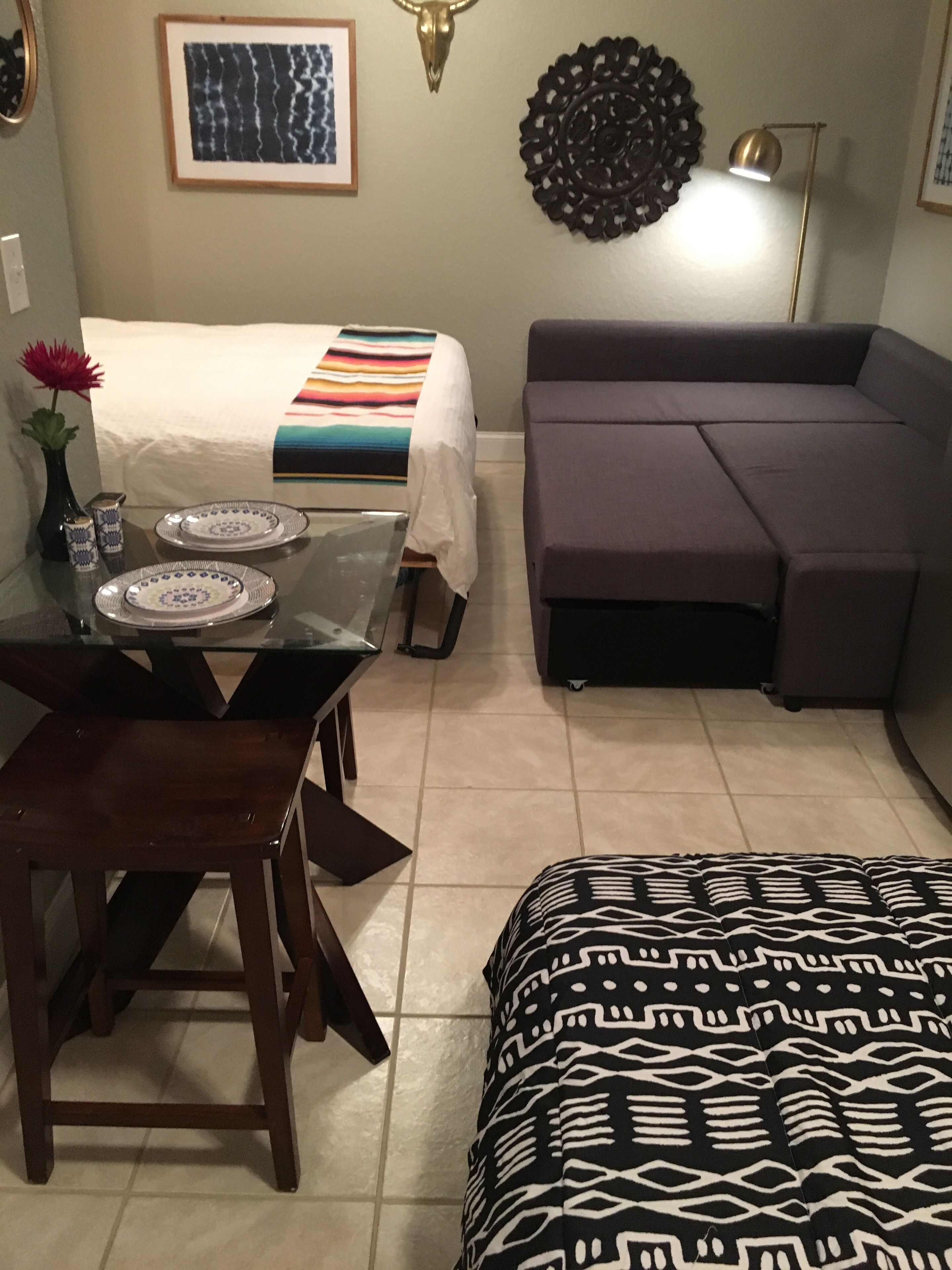 Ein Hotelzimmer mit einem Bett, einem Sofa, einem Esstisch mit Tellern, einer Vase mit einer Blume und einer Wanddekoration.