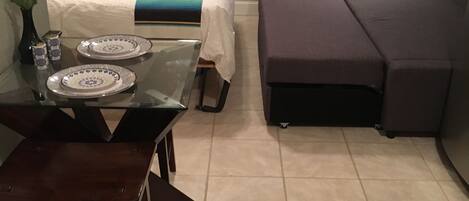 A hotel room with a bed, a sofa, a dining table with plates, a vase with a flower, and a wall decoration.