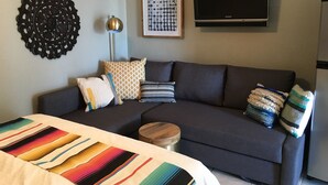 A modern living room with a dark grey sofa, a colorful striped bedspread, a round wooden side table, and a flat-screen TV mounted on the wall.