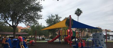 A playground with slides and climbing structures, surrounded by a fence and trees.