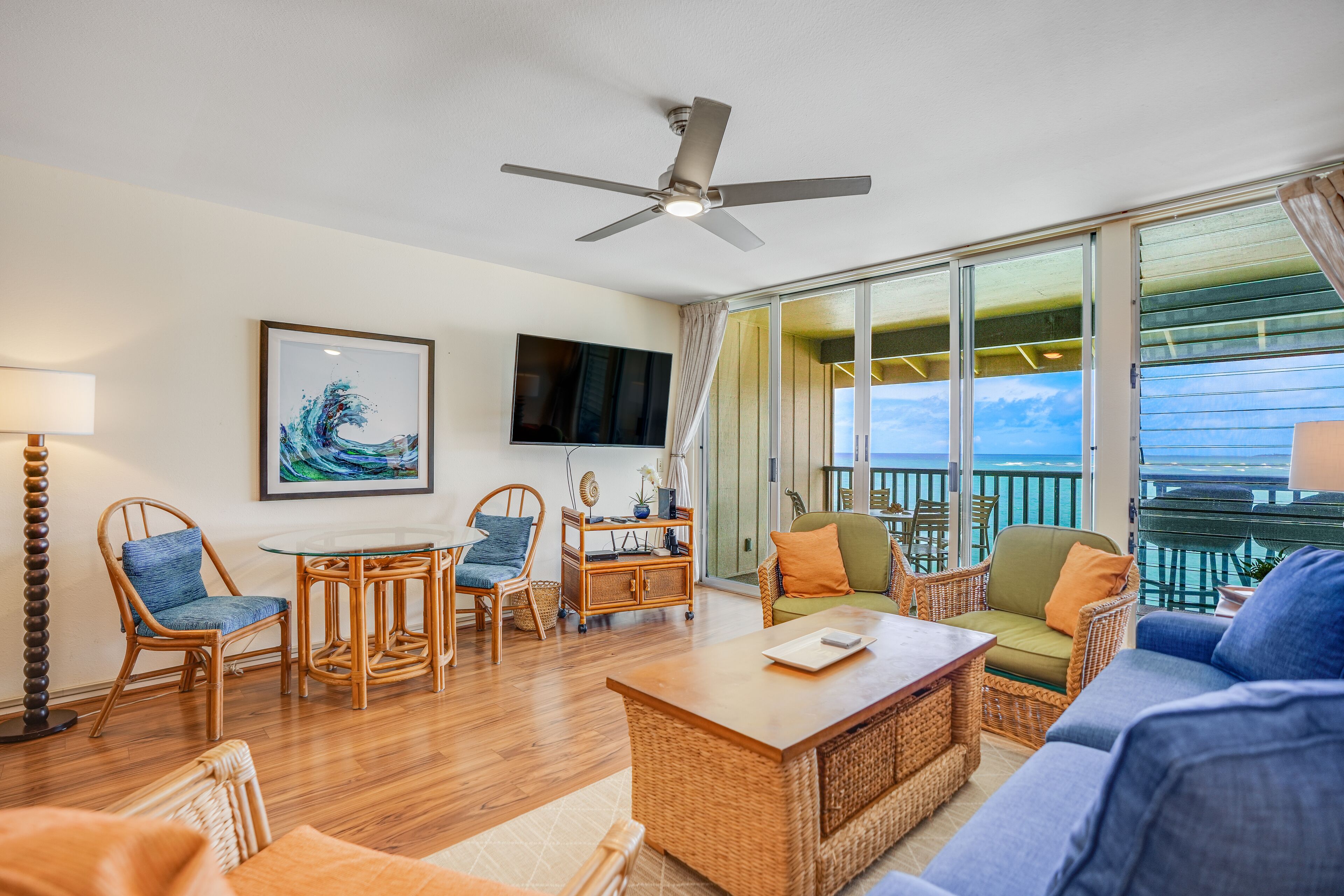 A spacious living room with a large window, wicker furniture, a ceiling fan, and a flat-screen TV.