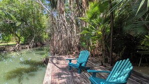 Eine Holzterrasse mit zwei türkisfarbenen Adirondack-Stühlen und einem Tisch, umgeben von dichter tropischer Vegetation und einem Gewässer.