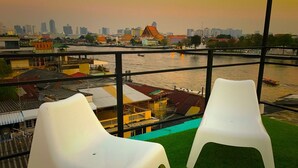 Two white plastic chairs on a balcony with a view of a river and a cityscape in the background. - Pho Hostel (Bangkok)