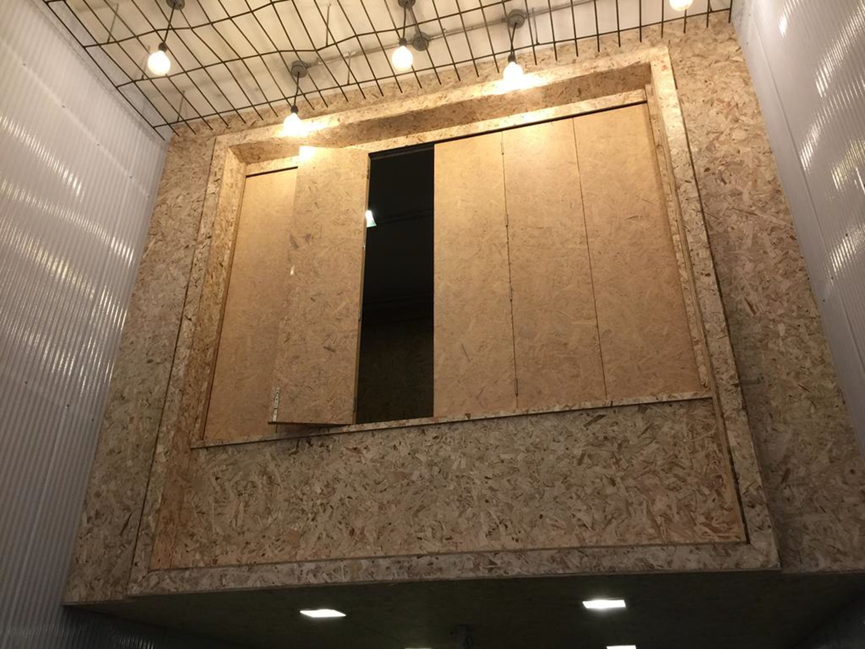 Una habitación con una pared grande y sin terminar, paneles de madera a la vista y una serie de luces en el techo.