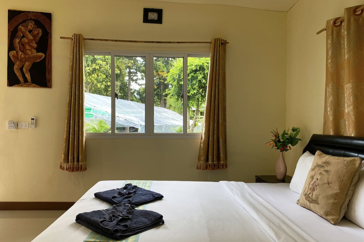 A hotel room with a bed, a window with curtains, a painting on the wall, a vase with flowers, and a thermostat on the wall.