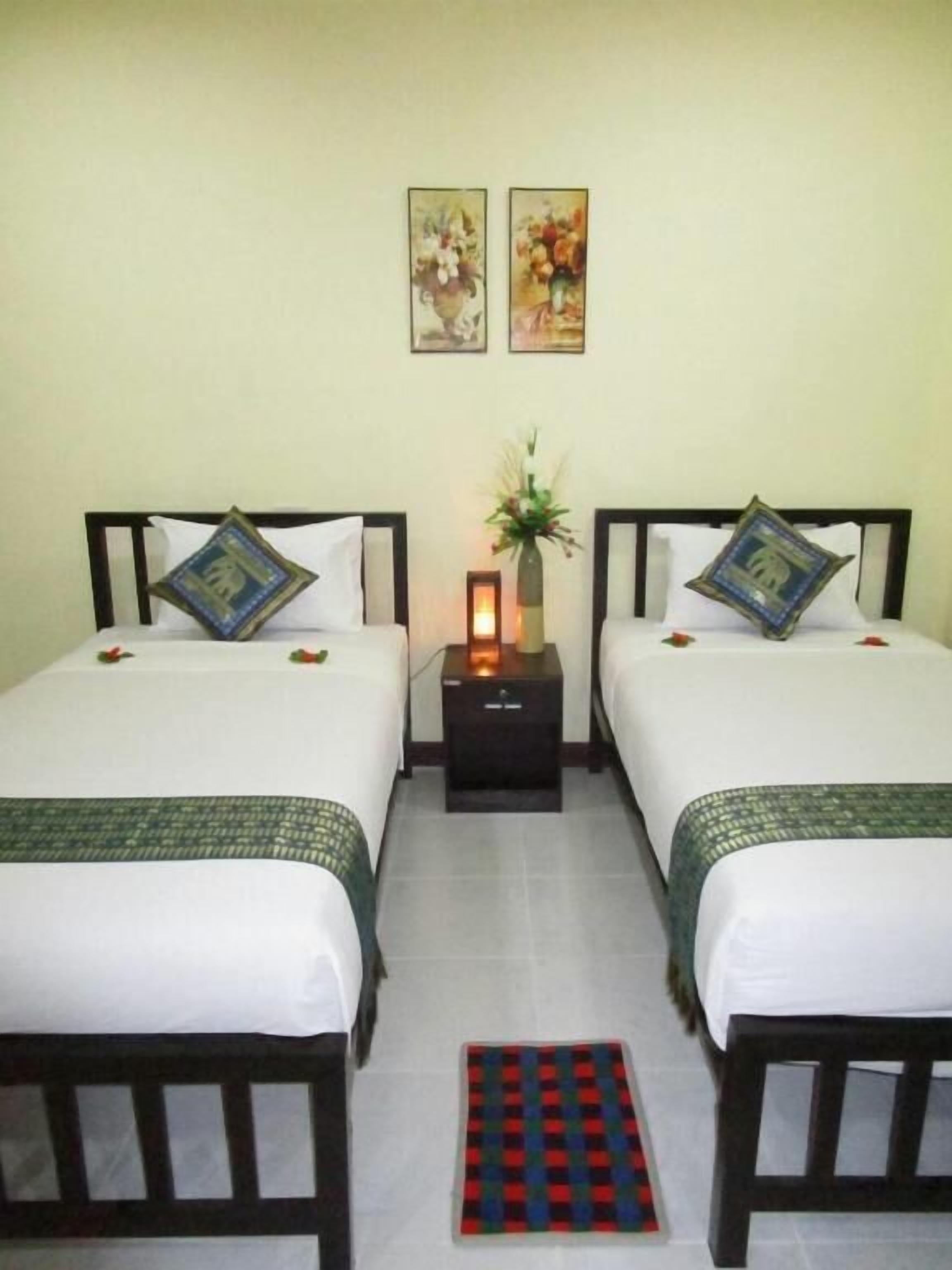 Habitación de hotel con dos camas, cada una con una almohada y una mesita con lámpara. Dos cuadros enmarcados adornan la pared sobre las camas.