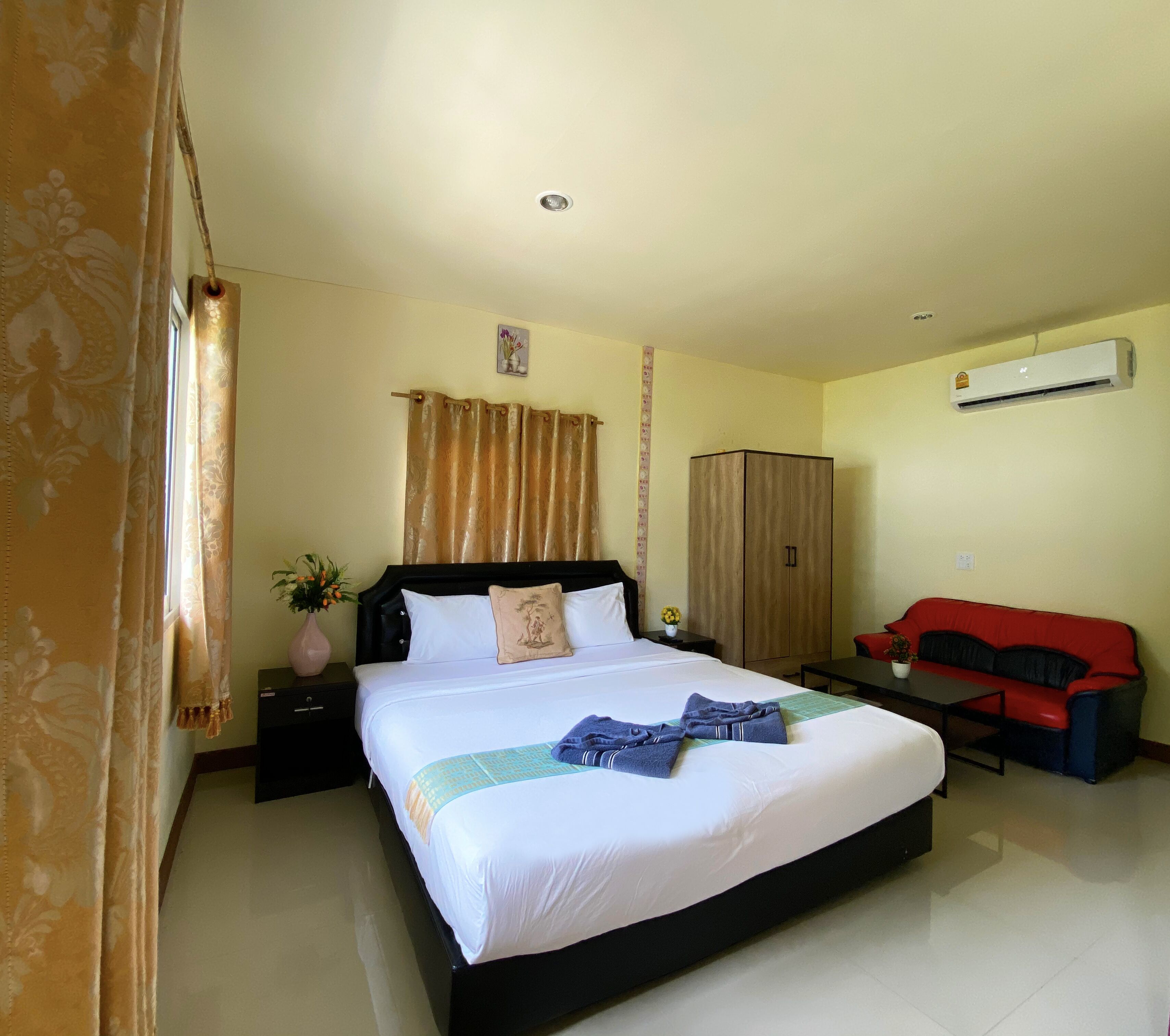 Habitación de hotel con una cama grande, un sofá rojo, un aire acondicionado y una mesita de noche con un florero.