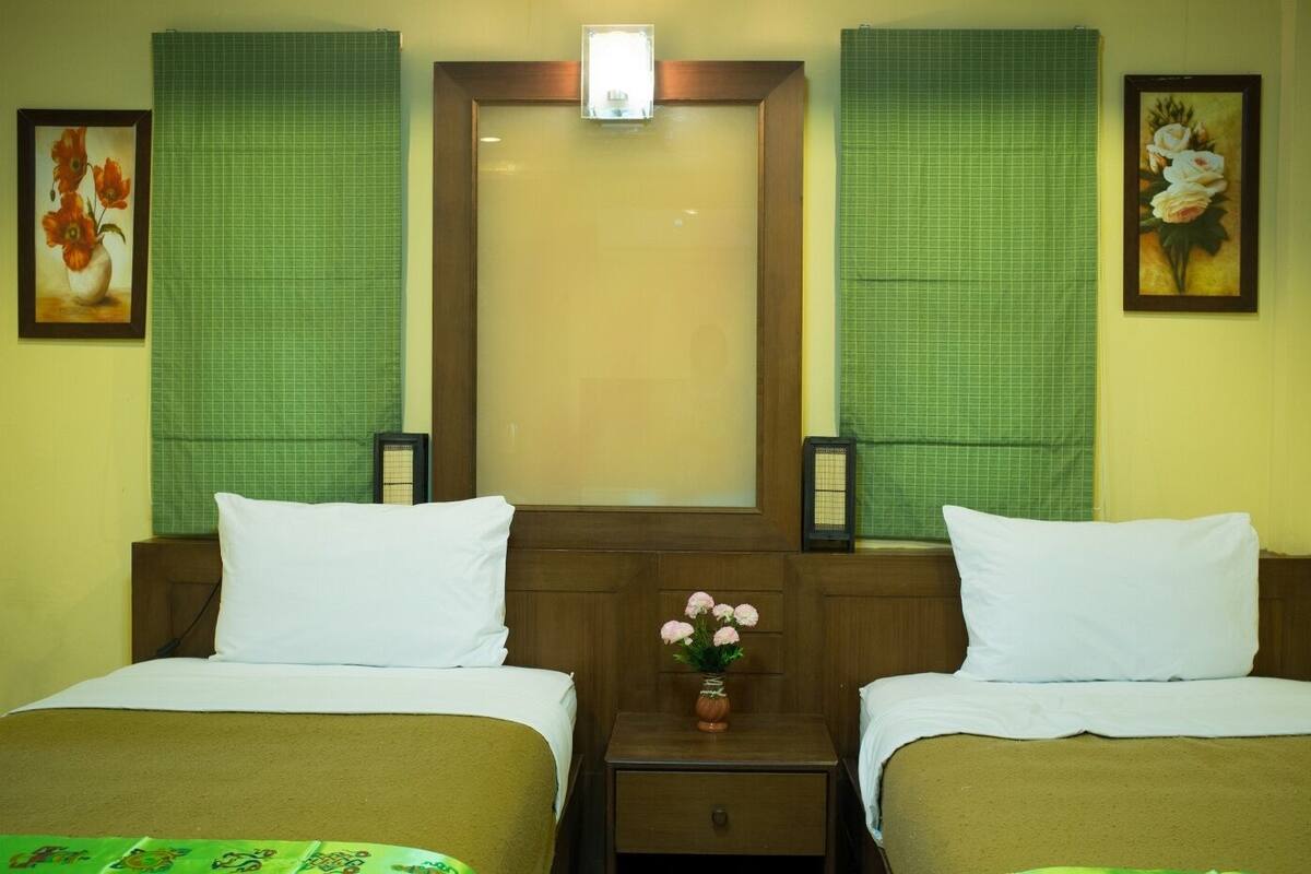 A hotel room with two beds, a nightstand with a vase of flowers, and green curtains.