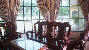 A room with ornate wooden furniture, large windows with patterned curtains, and a view of palm trees and a building outside. - 7 Makara Hotel (Kampong Cham)