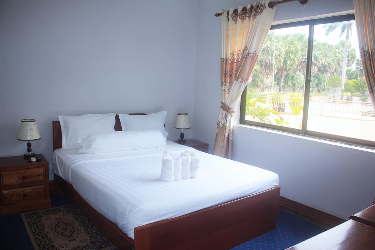 Una cama bien tendida con sábanas blancas, dos lámparas de noche, una mesita de madera y vistas a palmeras a través de una ventana con cortinas.