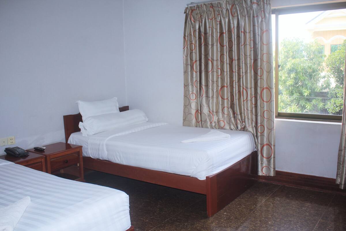 Una habitación de hotel con una cama individual, una mesita de noche de madera con un teléfono y una ventana con cortinas.