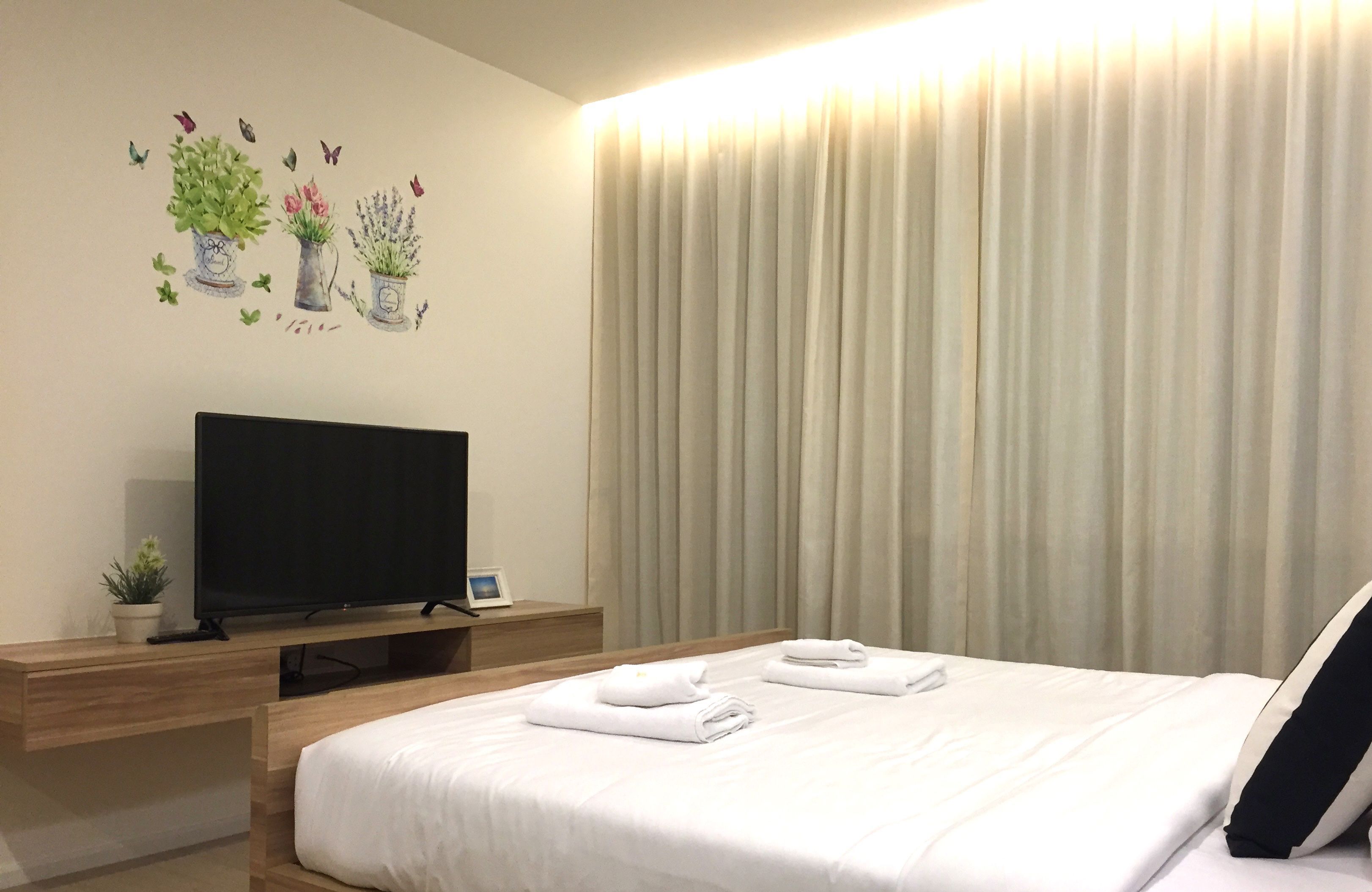 A hotel room with a bed, a flat-screen TV on a wooden console, a wall-mounted artwork of potted plants, and a large window with curtains.