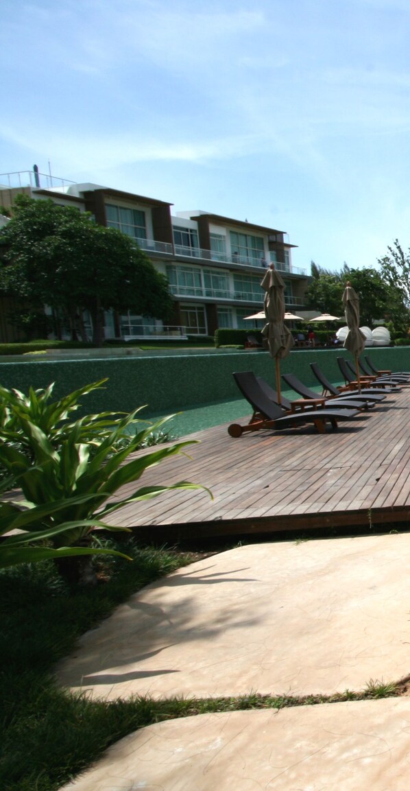 A poolside area with lounge chairs and umbrellas, a wooden deck, and a building with large windows in the background.