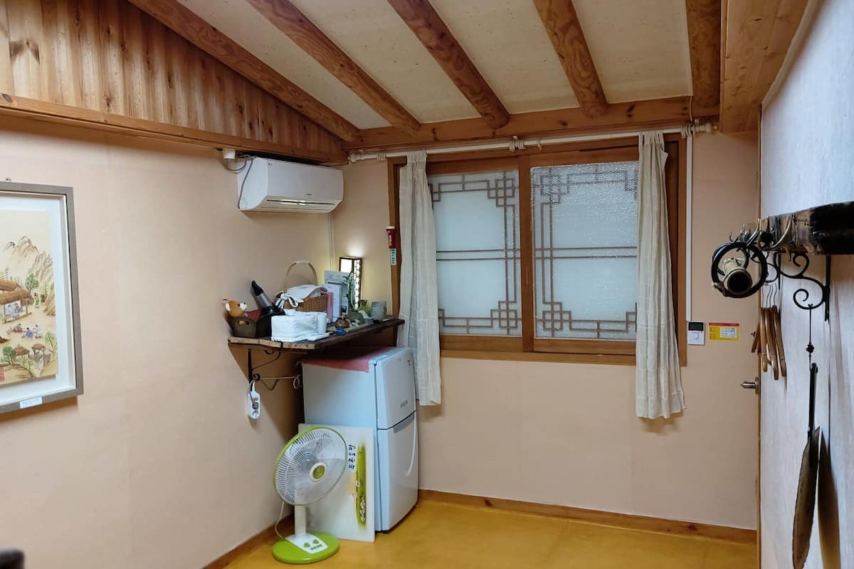 A small kitchen with a refrigerator, a fan, and a window with curtains.