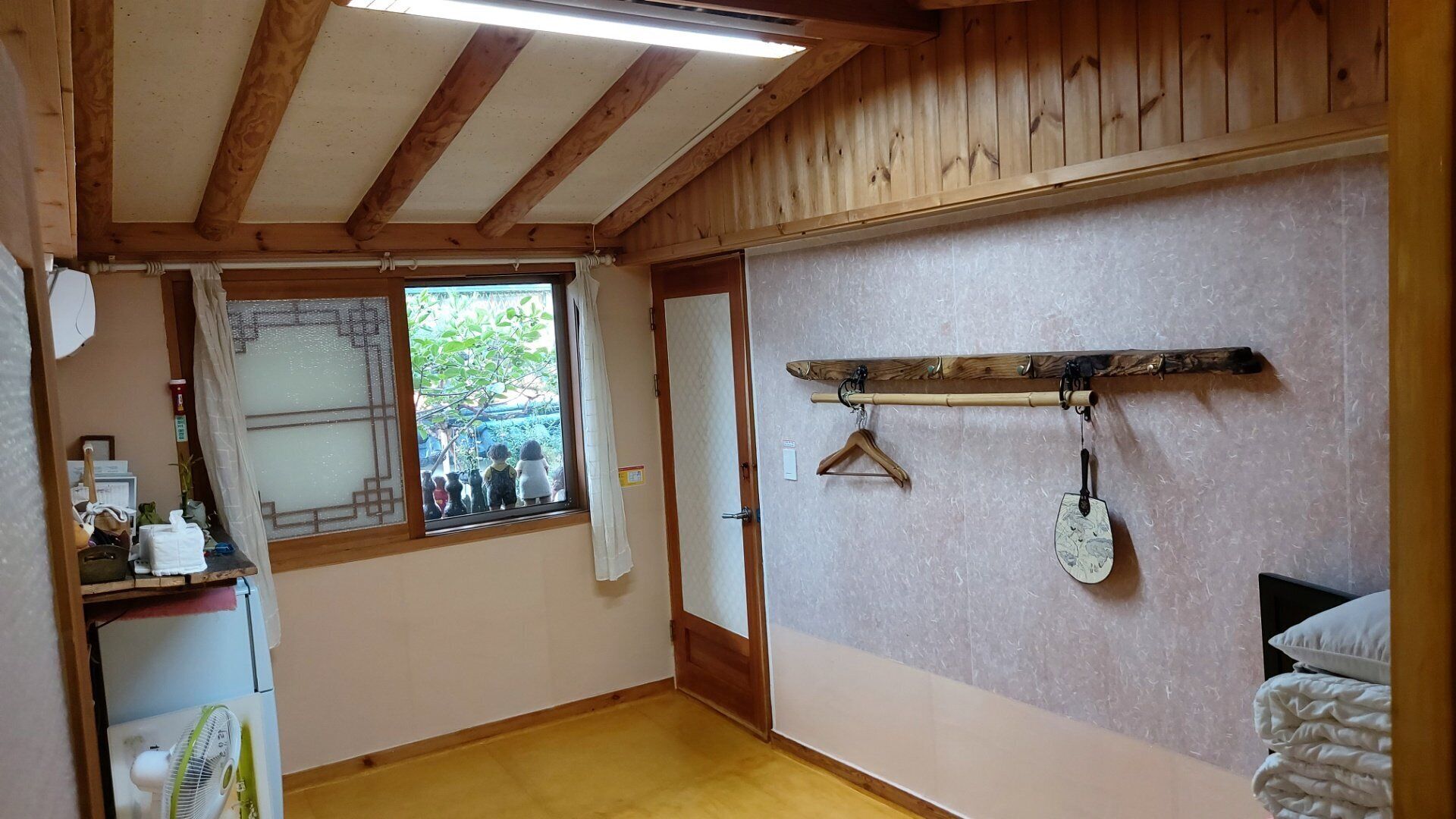 A room with wooden beams, a window with a view of greenery, a wooden shelf with a paddle, and a bed with a white comforter.