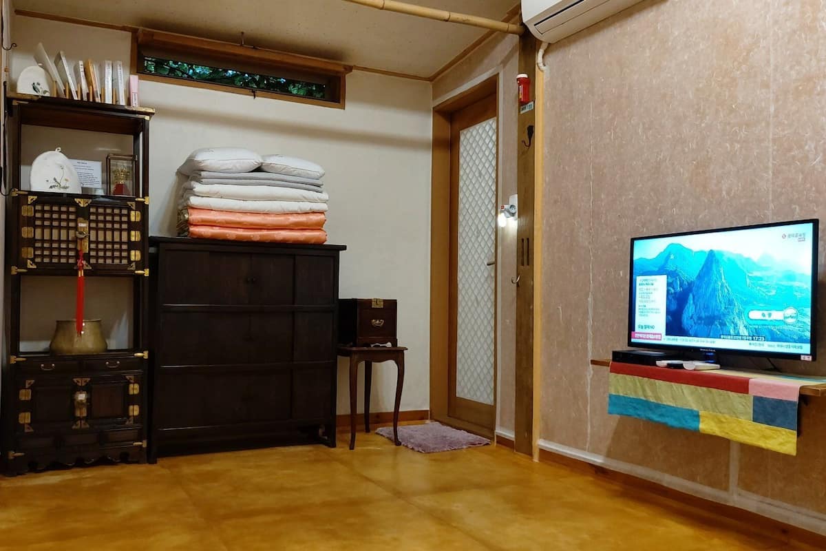 A room with a television on a stand, a wooden cabinet with folded blankets, and a small table with a lamp.