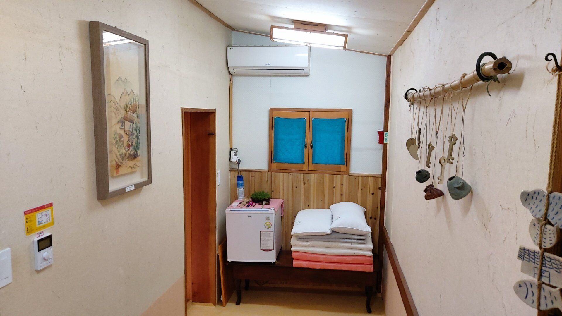 A compact room with a bed, a small table with a plant, a window with blue shutters, and a wall-mounted air conditioner.