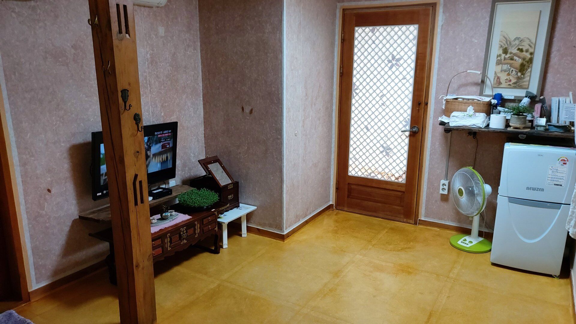 A compact living space with a wooden door, a small refrigerator, a television, and a fan.