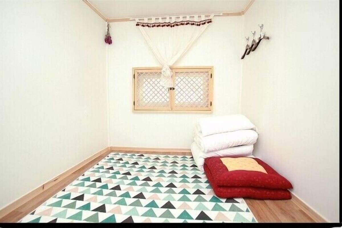 Een kleine, verzorgde kamer met een vloer met geometrisch patroon, een raam met houten kader en witte gordijnen, en een wandplank met decoratieve objecten.