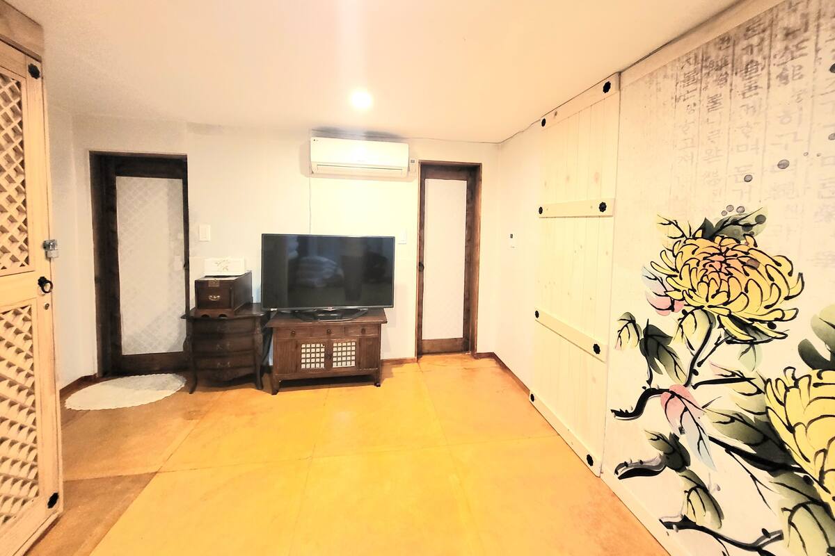 Une pièce avec un téléviseur posé sur un meuble en bois, un petit placard et un mur orné d’une fresque florale.