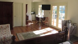 A spacious living room with a wooden floor, a floral sofa, a dining area with a table and chairs, a kitchen in the background, and a large window with a view. - Honeyeater Cottage (Yellingbo)
