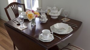 A dining table set with white porcelain tea set, including a teapot, cups, and saucers. There are also a vase with flowers and a glass on the table. - Honeyeater Cottage (Yellingbo)