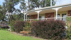 A house with a covered porch, brick walls, and a garden with a bench and various plants. - Honeyeater Cottage (Yellingbo)