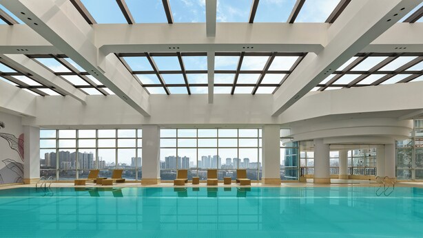 Une vaste piscine intérieure à l’eau d’un bleu limpide, entourée de chaises longues et dotée de grandes fenêtres offrant une vue sur le panorama urbain.