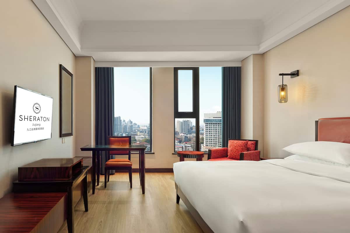 A hotel room with a bed, a desk, a chair, and a TV displaying the Sheraton logo.