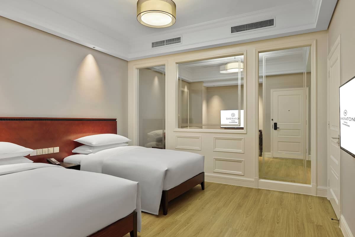 A hotel room with two single beds, a wooden headboard, a large mirror, a wardrobe, and a ceiling light.