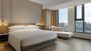 Une chambre d’hôtel moderne avec un grand lit, une table de chevet avec un téléphone, un banc et une vue sur la ville à travers de grandes fenêtres.