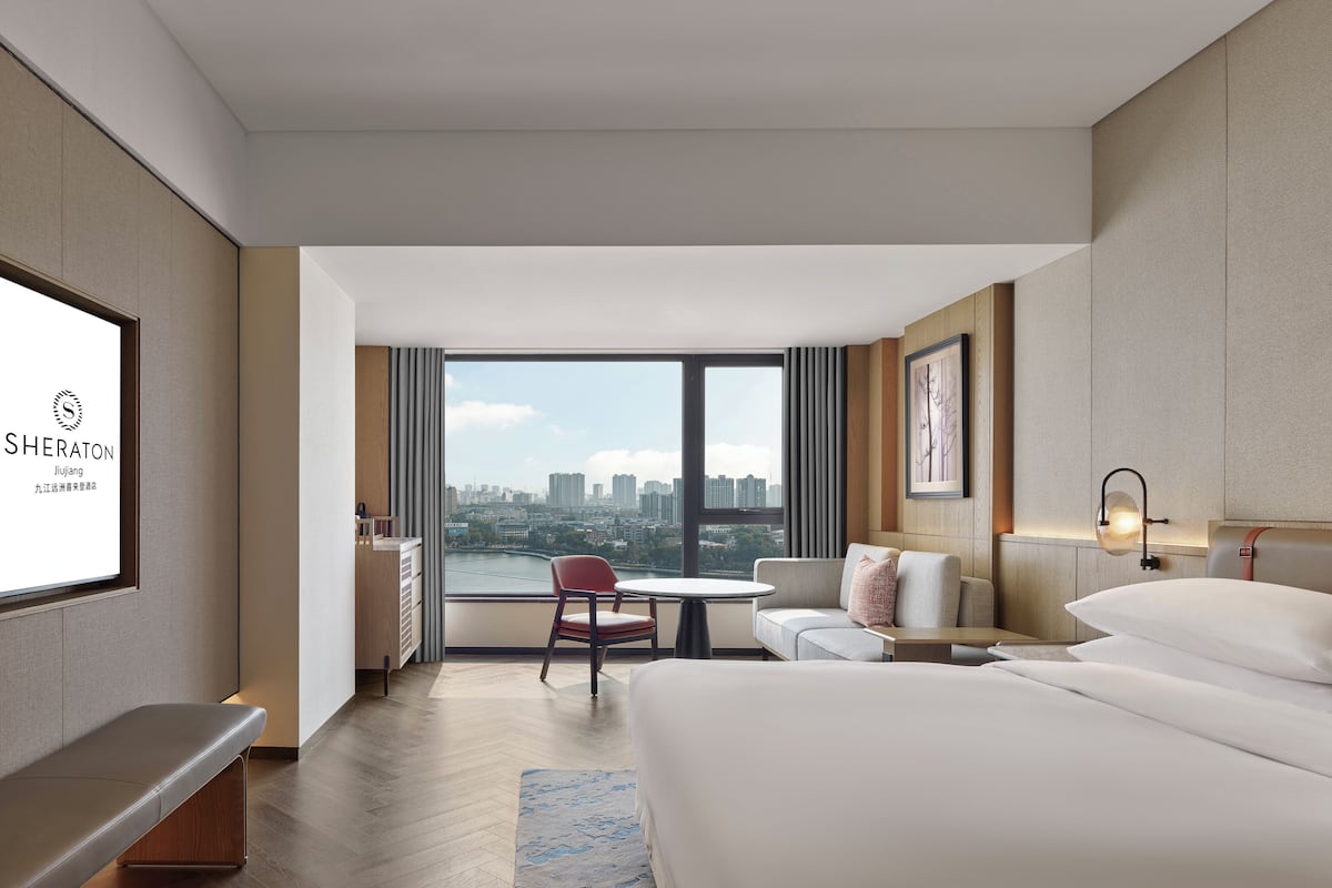 A modern hotel room with a large bed, a seating area with a table, a TV screen displaying hotel branding, and a city view through large windows.