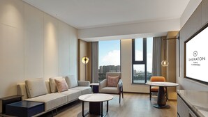 Une chambre d’hôtel moderne, dotée d’un canapé, de fauteuils, d’une table basse et d’une table à manger. De grandes fenêtres offrent une vue sur la ville.