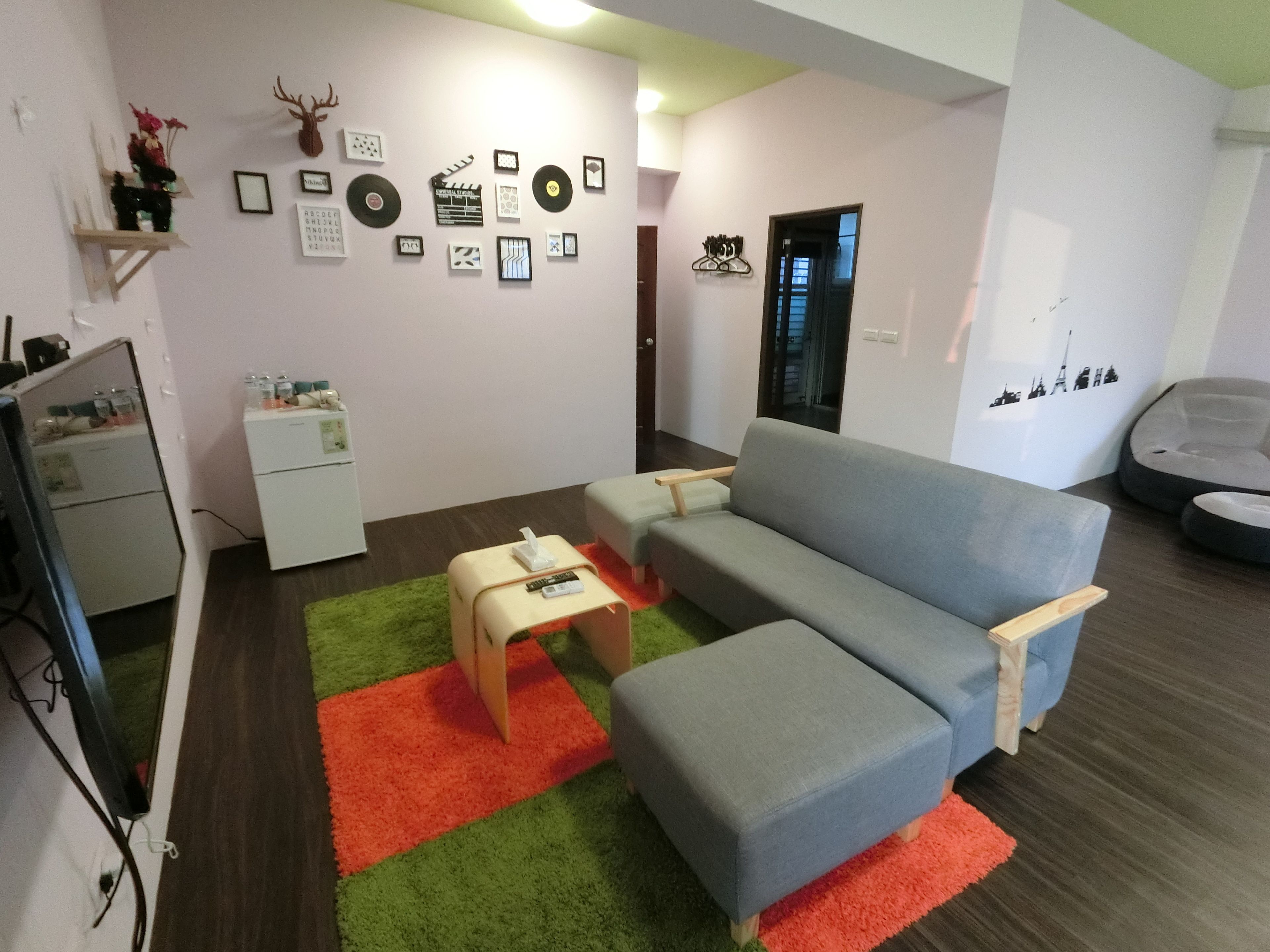 Ein modernes Wohnzimmer mit einem grauen Sofa, einem kleinen Tisch und einem Teppich in Grün und Rot. Im Hintergrund sind Wanddekorationen und eine Tür zu sehen.