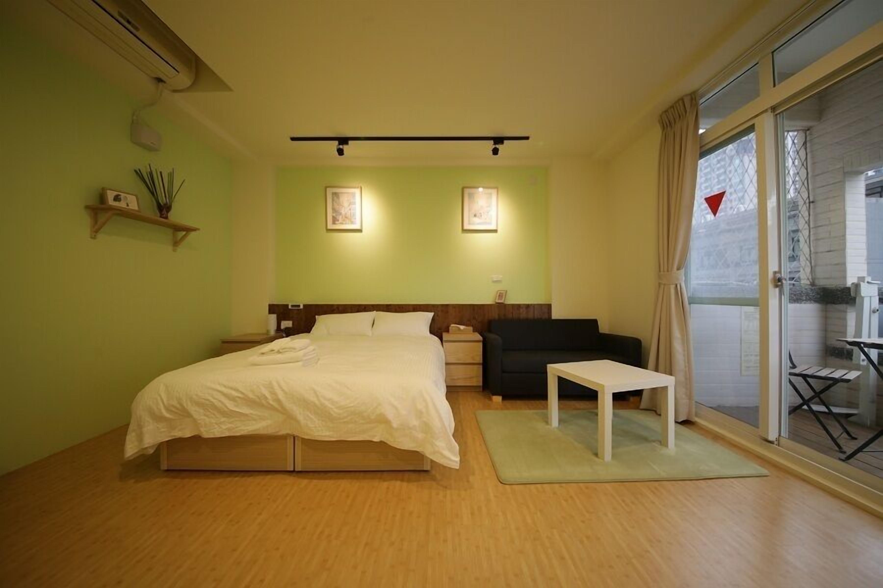 A bedroom with a bed, bedside table, a small table, a sofa, and a sliding glass door leading to an outdoor area.