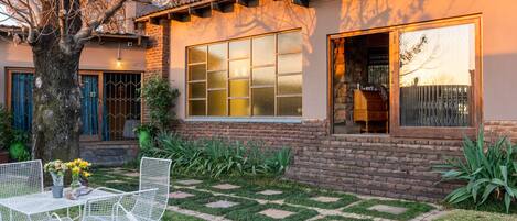 A house with a brick exterior, a large window with multiple panes, a patio with a table and chairs, and a tree in the foreground.
